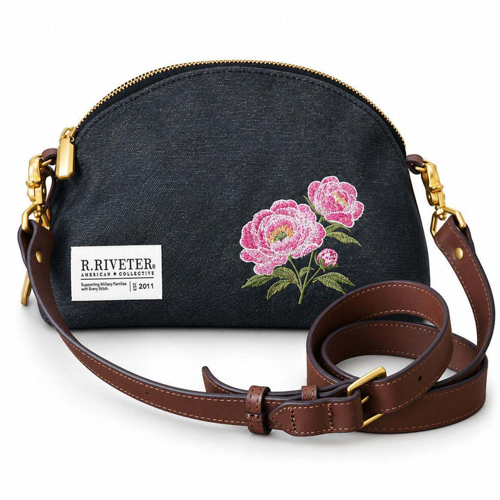 A black canvas crossbody bag with a brown leather strap, gold-tone hardware, and pink floral embroidery on the front. A white brand label reading 'R.Riveter'