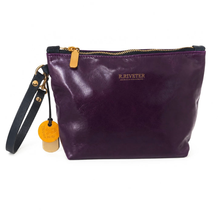 A purple leather zippered pouch bag by R. Riveter, featuring a black wristlet strap with gold-tone hardware and a round tan leather hangtag, shown on a white background.