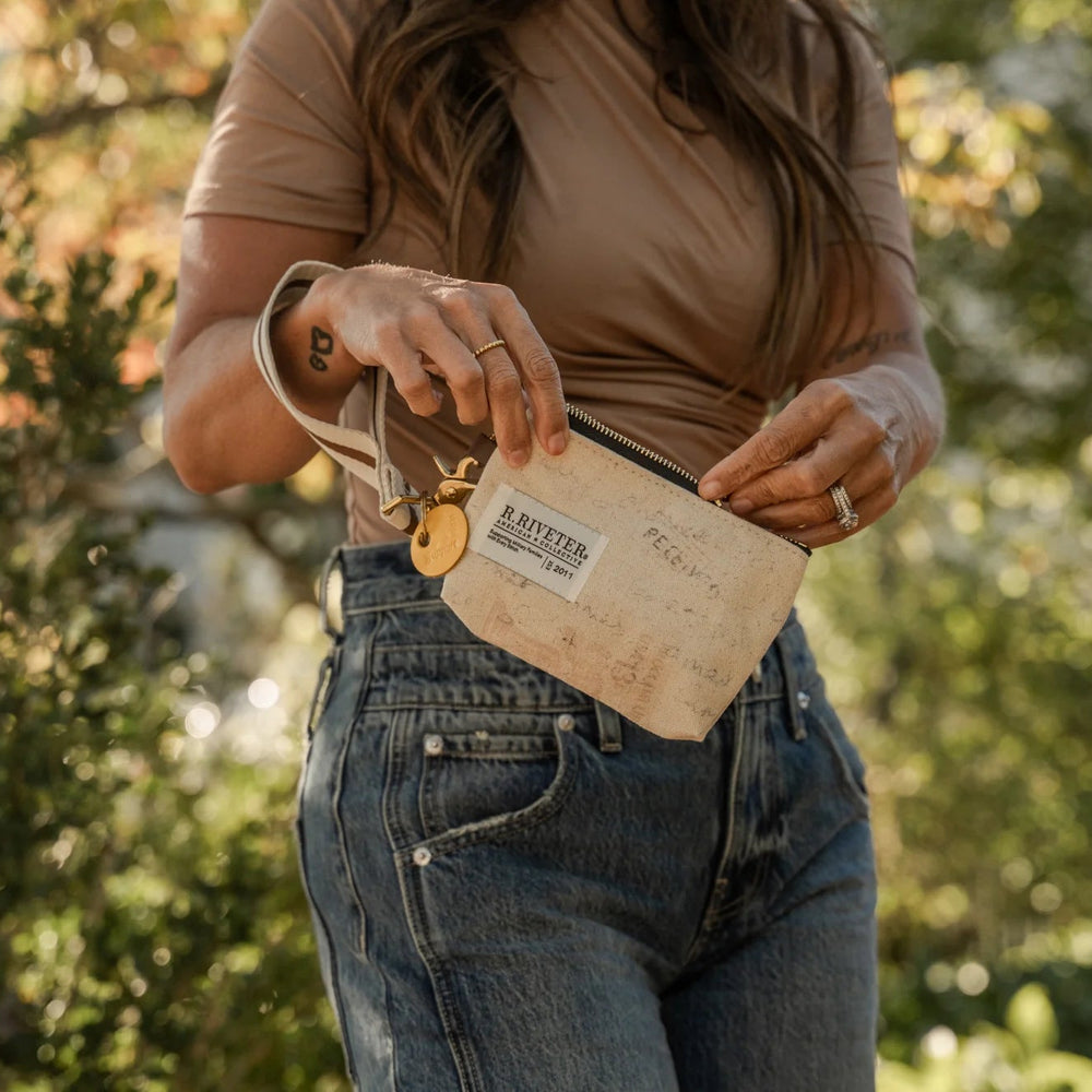 A woman outdoors wearing a brown short-sleeve top and blue jeans, holding a beige canvas pouch with a leather strap and gold-tone hardware.