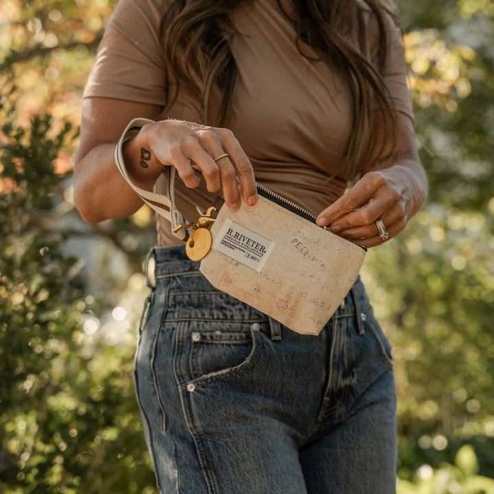 A woman outdoors wearing a brown short-sleeve top and blue jeans, holding a beige canvas pouch with a leather strap and gold-tone hardware.