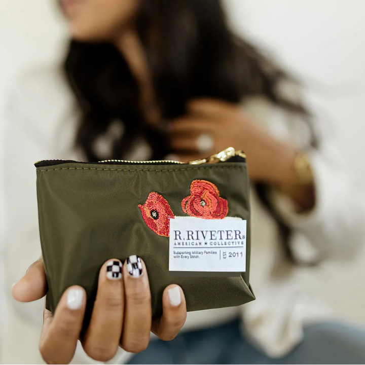 A woman holds a small olive green zip pouch with red poppy embroidery and a white R.Riveter label.