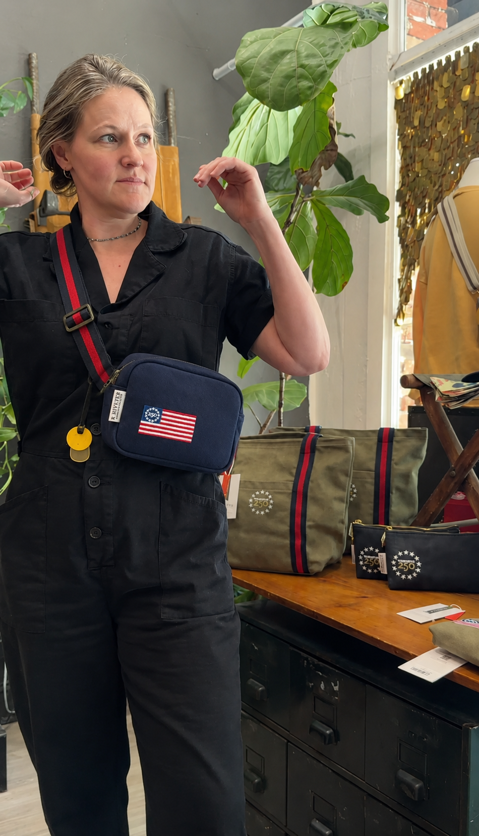 Person wearing a black outfit with a blue bag featuring an American flag, standing in a room with plants and furniture.