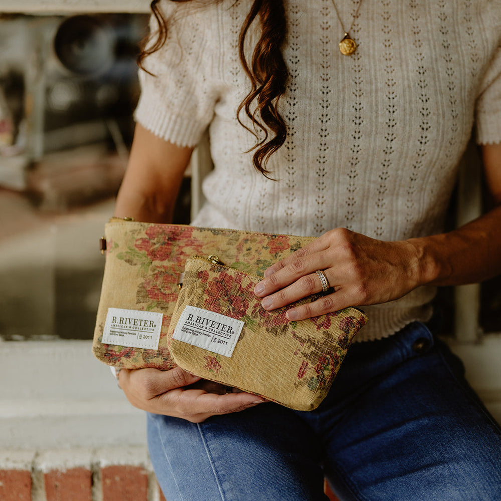 A woman in a cream knit sweater and blue jeans holds a small floral pouch with two visible 'R.RIVETER' labels.