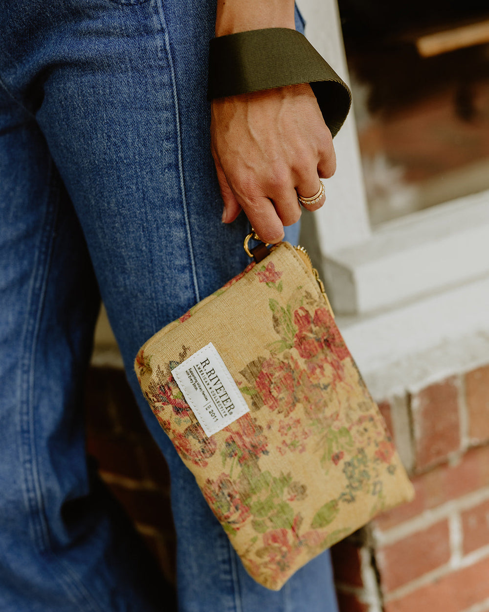 A person wearing blue jeans holds a small floral clutch bag with a white label. The clutch has a vintage-style pink and green floral print on a tan background, and the person is also wearing an olive green wide strap accessory.