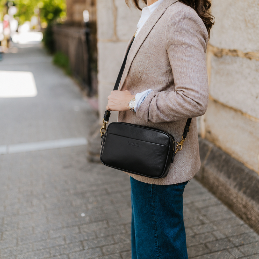 A woman stands on a stone sidewalk beside a stone wall, wearing a light beige blazer, blue jeans, and carrying a black crossbody bag with gold-tone hardware.