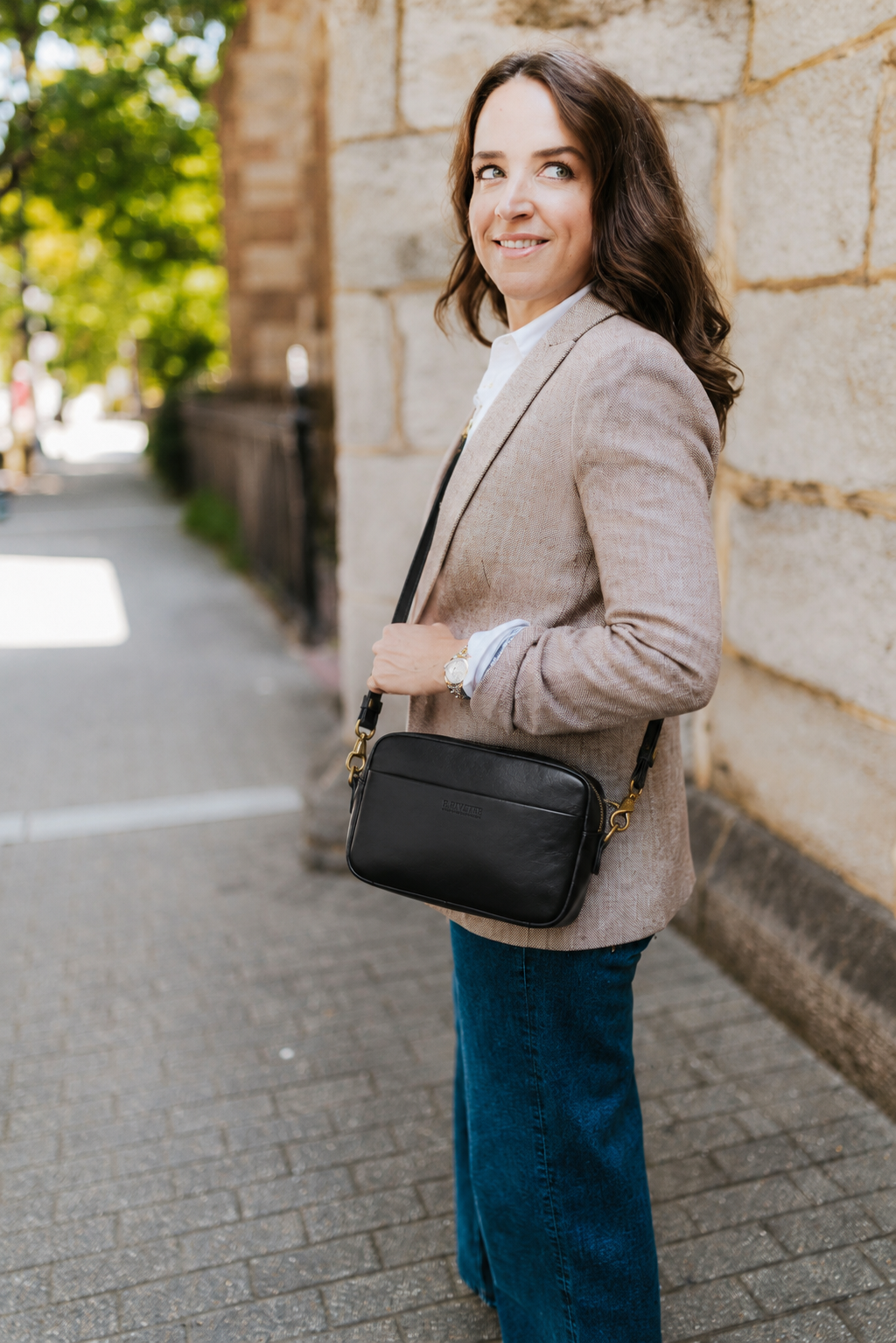 A woman stands on a stone sidewalk beside a stone wall, wearing a light beige blazer, blue jeans, and carrying a black crossbody bag with gold-tone hardware.