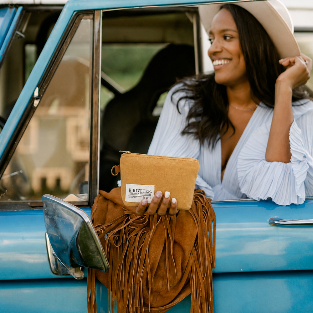Woman sitting in a vintage blue car holding a yellow canvas zipper pouch with a visible R.Riveter label.