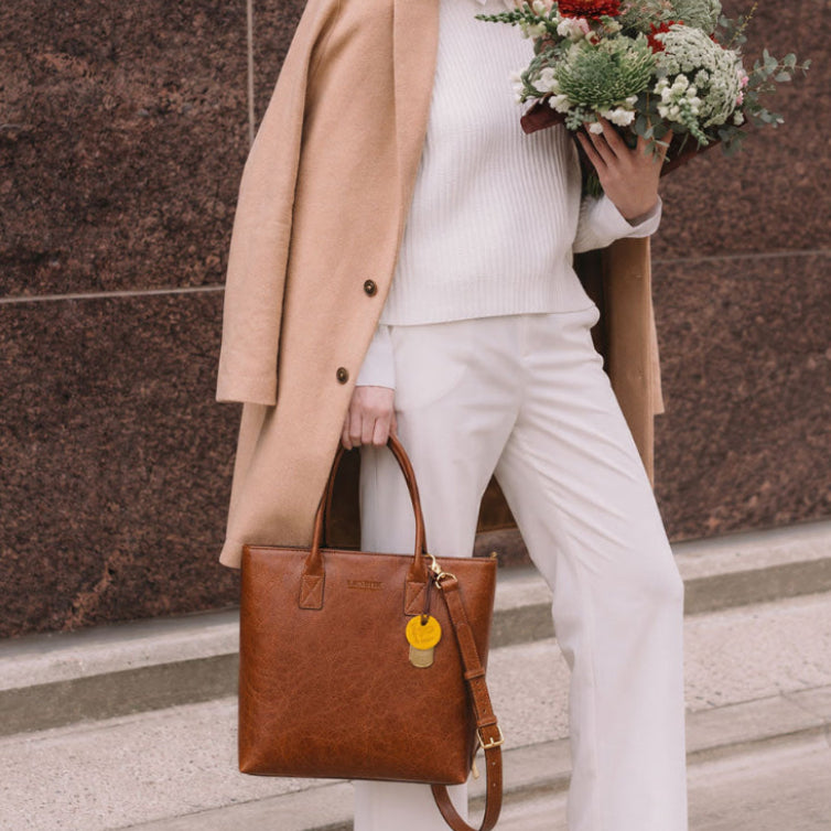 A woman stands outdoors against a brown stone wall, wearing a beige blazer, white ribbed sweater, and white wide-leg pants while holding a brown leather tote bag and a bouquet of flowers.