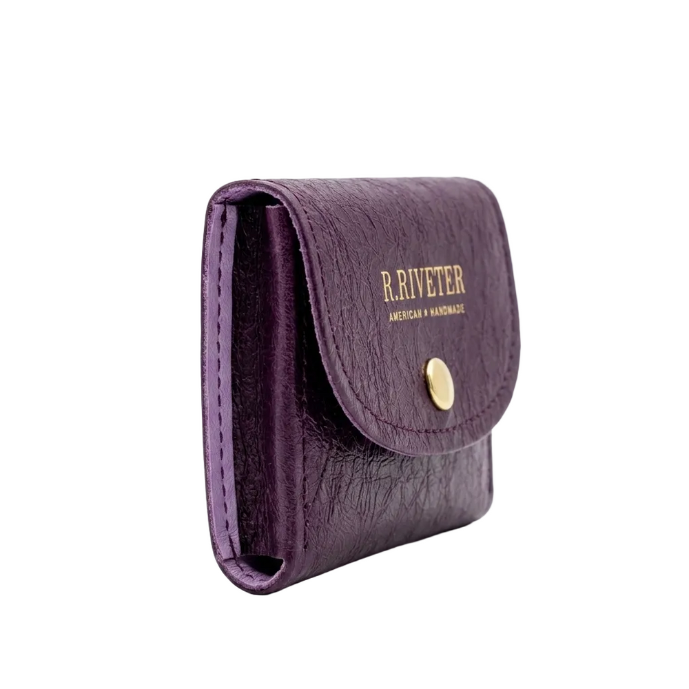 A small purple leather wallet with a flap closure, gold snap button, and gold 'R. RIVETER AMERICAN HANDMADE' branding on the front, shown on a clean white background.