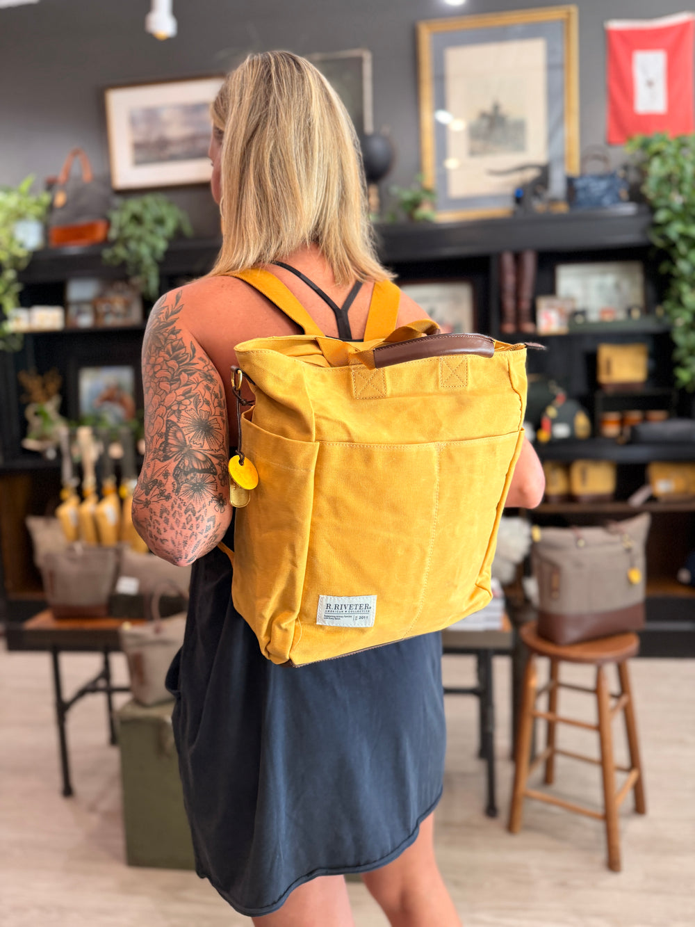 A woman viewed from behind wearing a mustard yellow canvas backpack with brown leather trim and a small white brand patch on the front, standing inside a rustic shop with wooden shelves and framed art in the background.