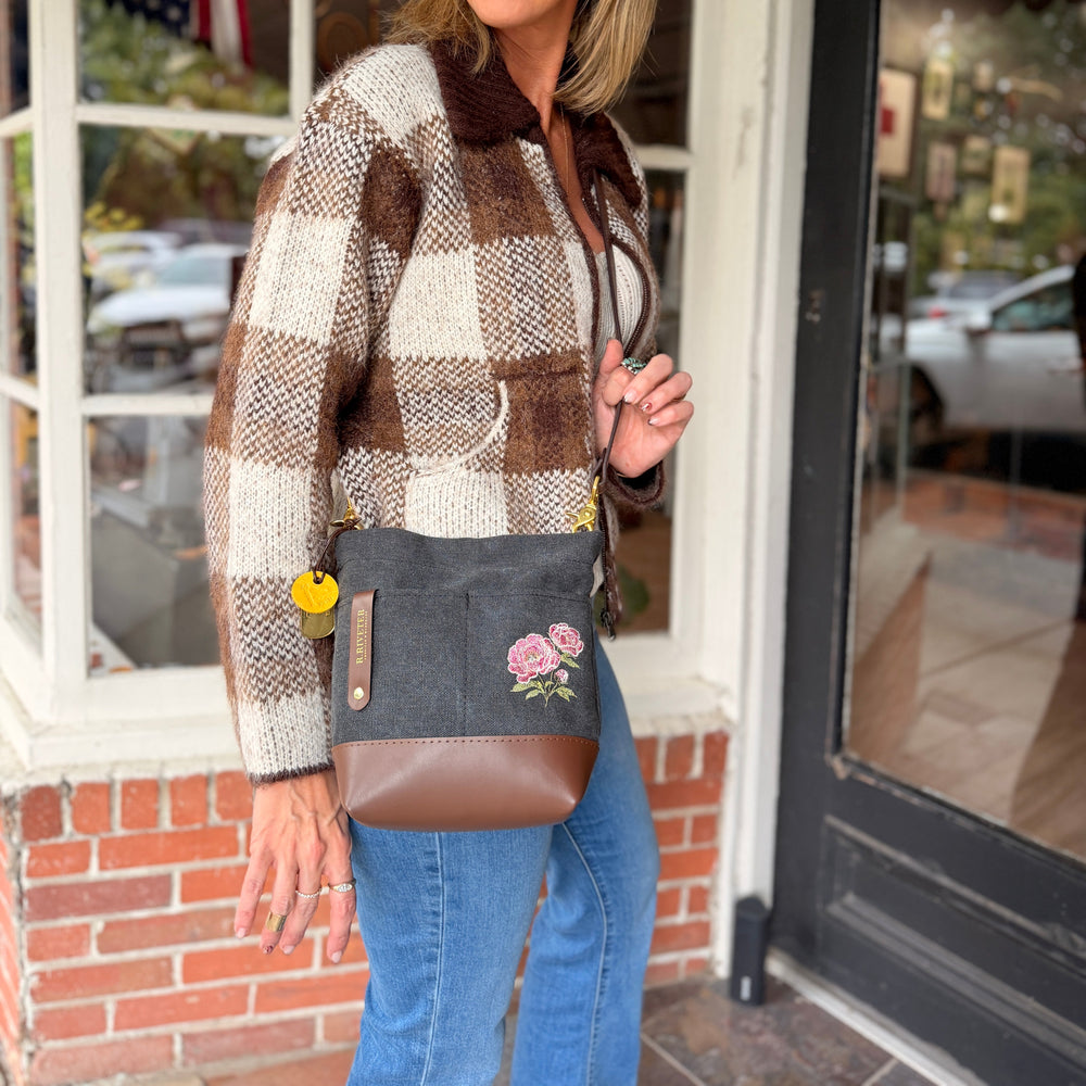 A woman wearing a brown and cream plaid flannel shirt jacket and blue jeans stands in front of a storefront window, carrying a black crossbody bag with brown leather trim and pink floral peony embroidery.