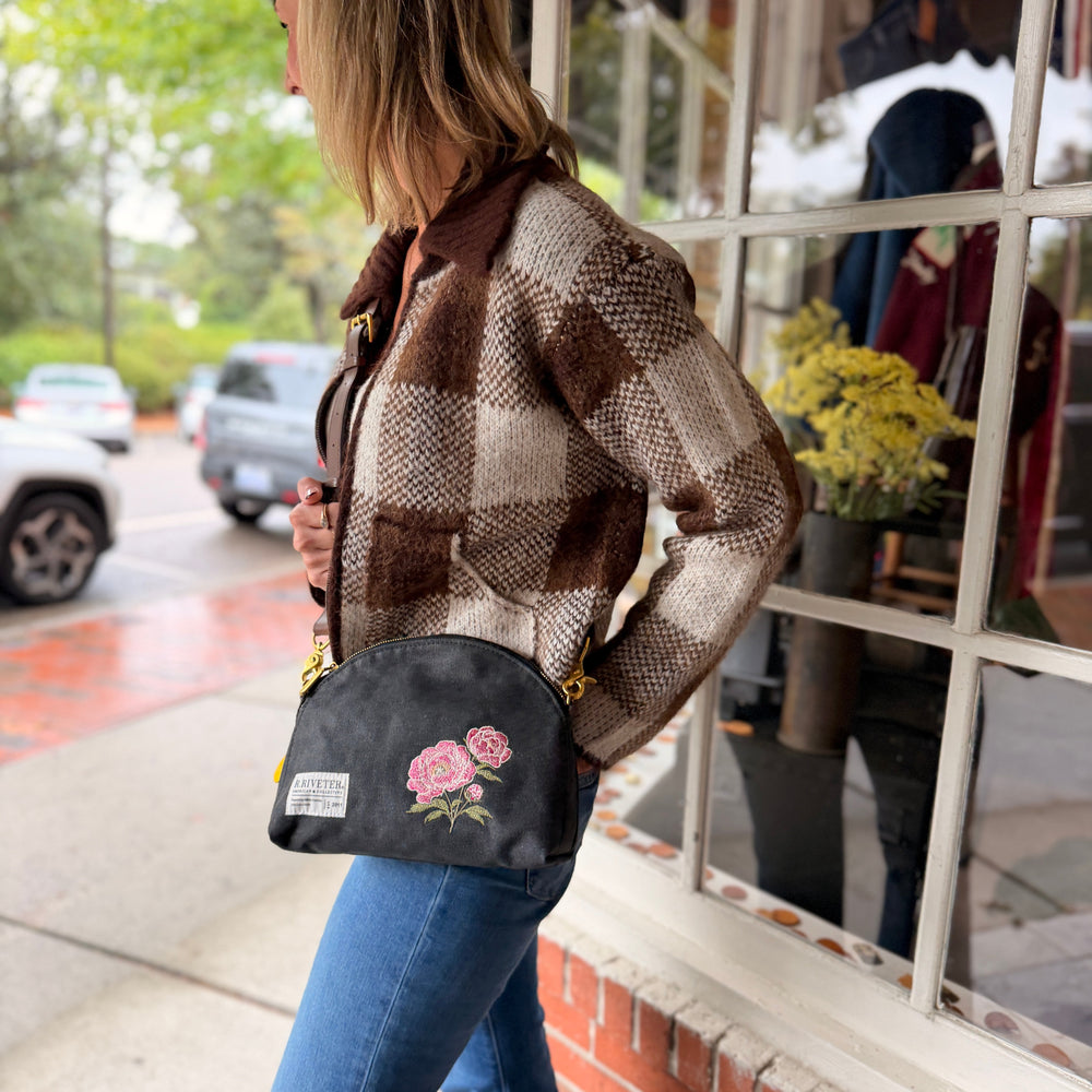 A woman wearing a brown and beige plaid jacket and blue jeans stands on a sidewalk outside a shop window, carrying a black crescent-shaped crossbody bag with pink peony embroidery and gold hardware.