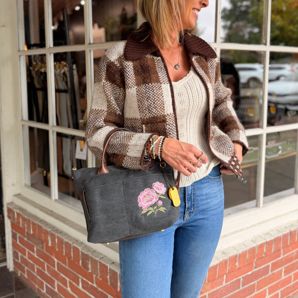 A woman stands in front of a shop window wearing a brown and beige plaid jacket, a cream ribbed top, and blue jeans, while carrying a dark gray tote bag with pink floral embroidery.