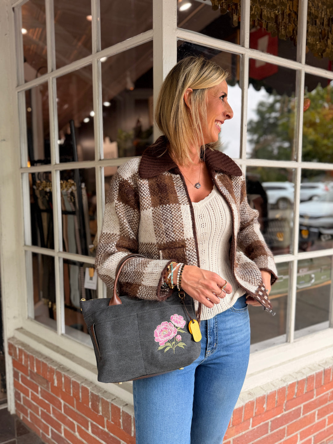 A woman stands in front of a shop window wearing a brown and beige plaid jacket, a cream ribbed top, and blue jeans, while carrying a dark gray tote bag with pink floral embroidery.