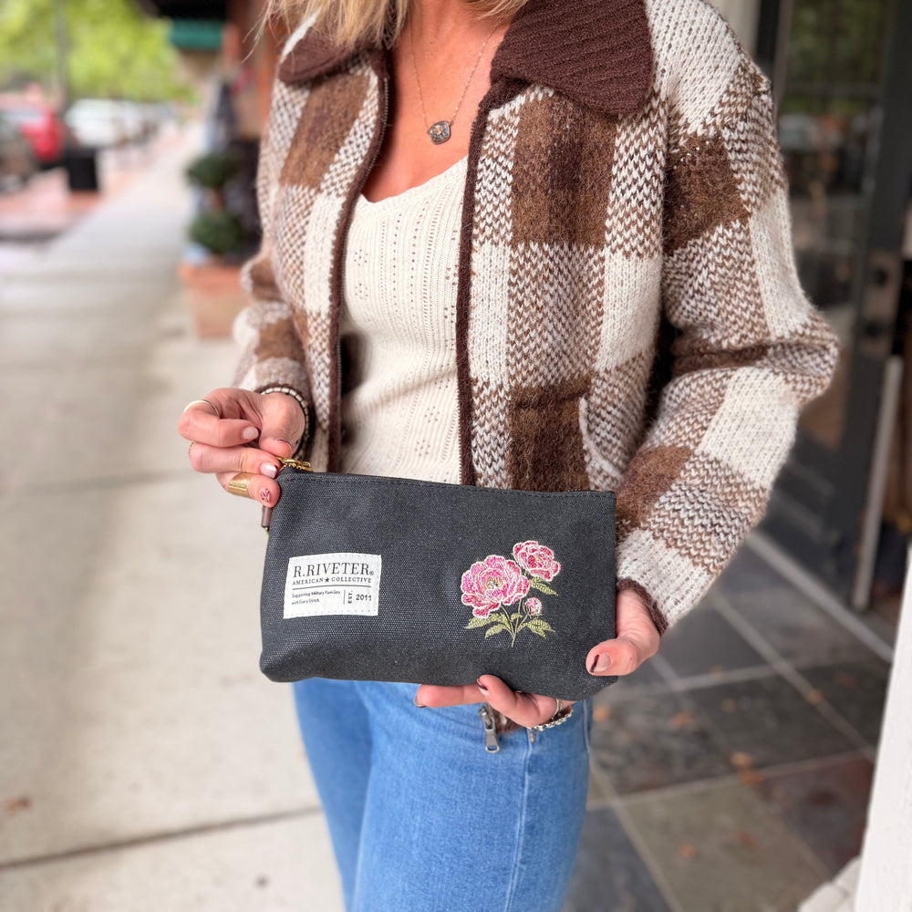 A woman wearing a brown and cream plaid knit jacket and blue jeans holds up a black canvas pouch with pink peony embroidery and a white 'R.RIVETER' brand label.