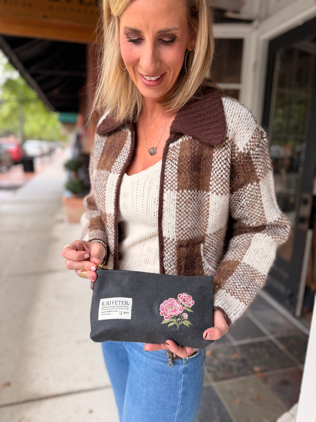 A woman wearing a brown and cream plaid knit jacket and blue jeans holds up a dark gray fabric pouch with pink rose embroidery and a white 'B. RITTEER' brand label.