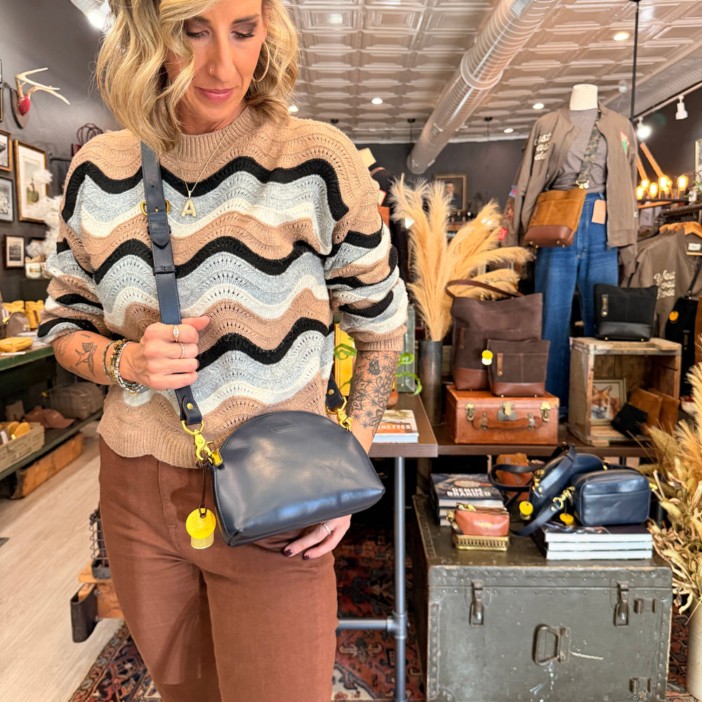 A woman wearing a beige, brown, and black wavy knit sweater and brown pants models a dark blue leather crossbody bag with yellow charms in a vintage-style boutique setting.