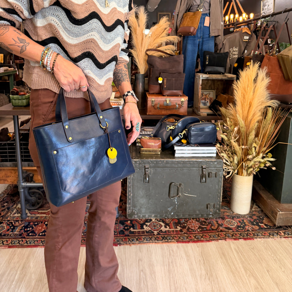 A woman in a cozy, patterned knit sweater and brown pants holds up a navy blue leather tote bag with gold hardware in a rustic, vintage-filled boutique.