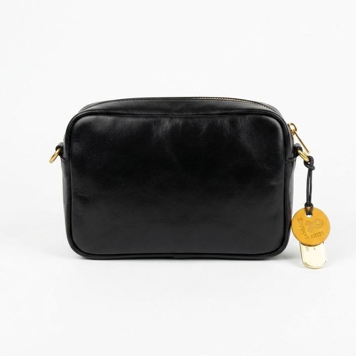 A black leather camera bag with gold-tone hardware and a hanging round gold tag, photographed against a clean white background.