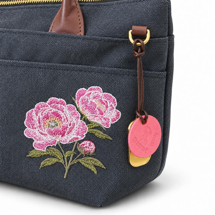 A close-up of a black washed canvas zippered pouch featuring embroidered pink peonies with green leaves on the front pocket. The bag has brown leather trim, gold-tone hardware, and a pink leather hangtag with a gold logo.