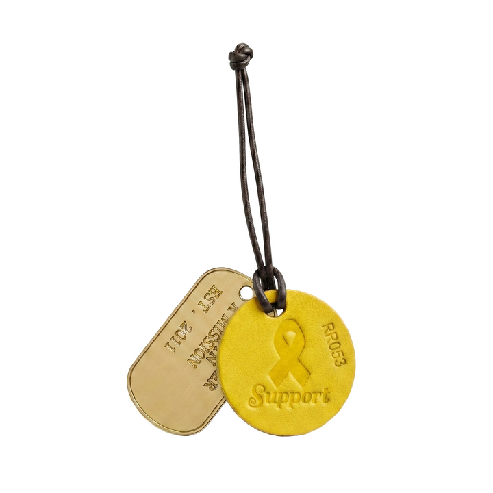 A round leather yellow support tag with a ribbon symbol, and a brass military dog tag on a white background.