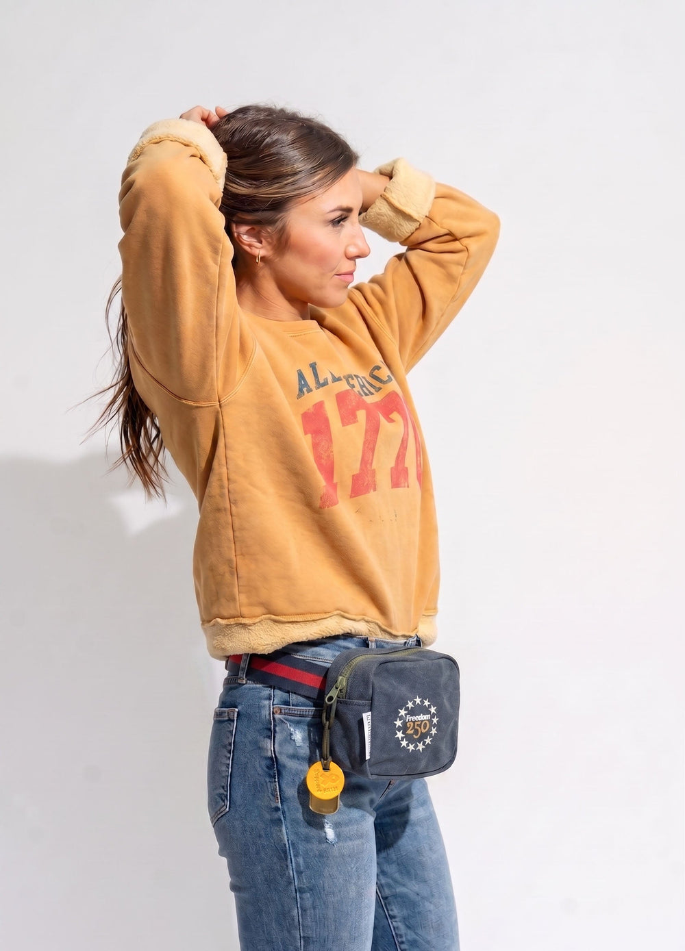 Person wearing a mustard yellow sweatshirt with red text and blue jeans, carrying a gray waist bag.