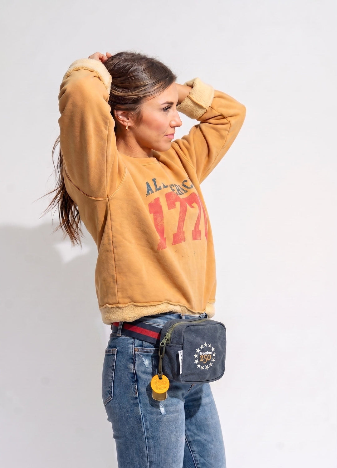 Person wearing a mustard yellow sweatshirt with red text and blue jeans, carrying a gray waist bag.