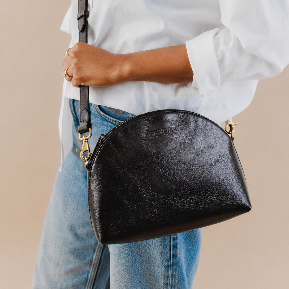 A person wearing a white button-up shirt and blue jeans carries a black faux-leather crescent-shaped shoulder bag with gold-tone hardware and a black strap against a plain beige background.