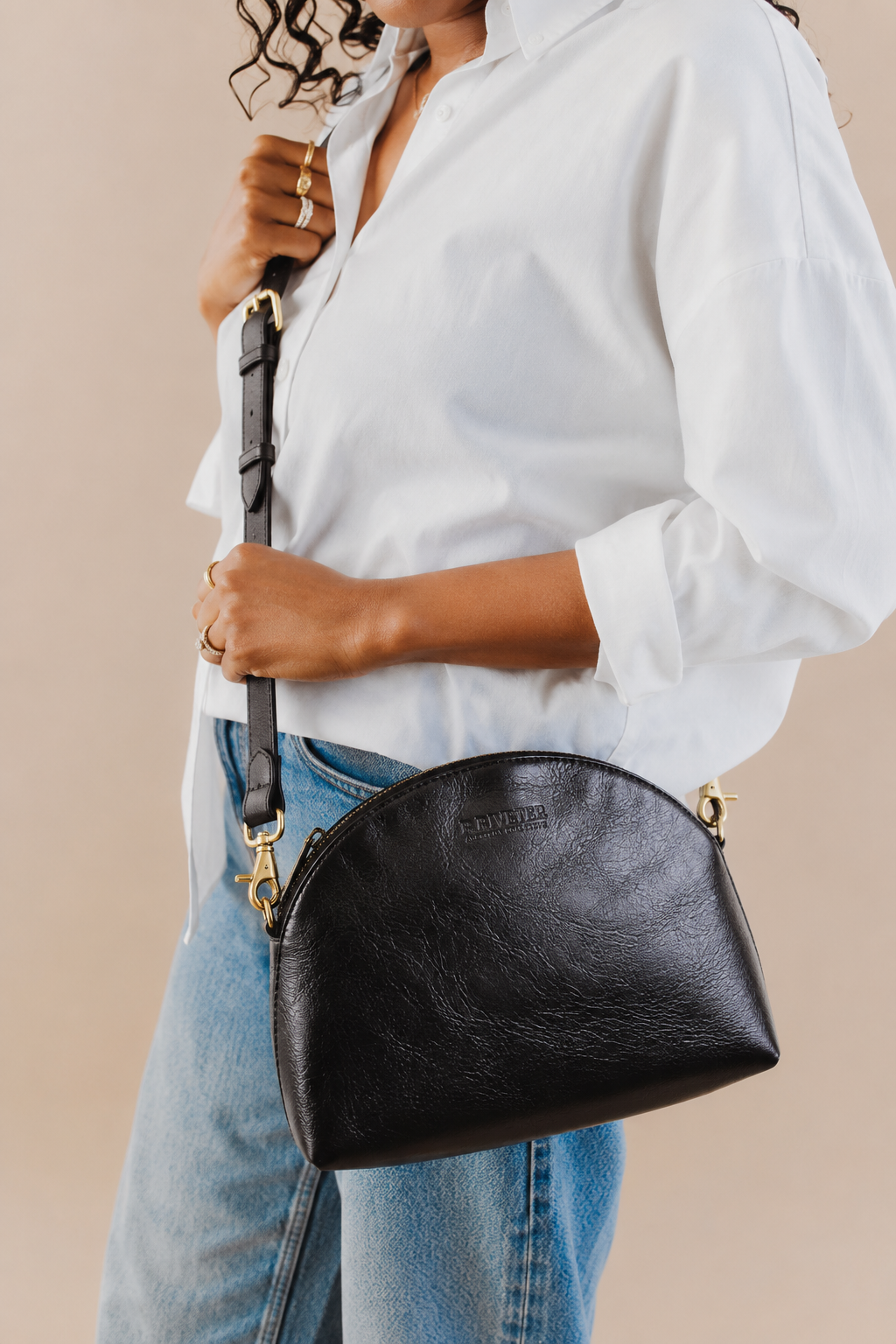 A person wearing a white button-up shirt and blue jeans carries a black faux-leather crescent-shaped shoulder bag with gold-tone hardware and a black strap against a plain beige background.