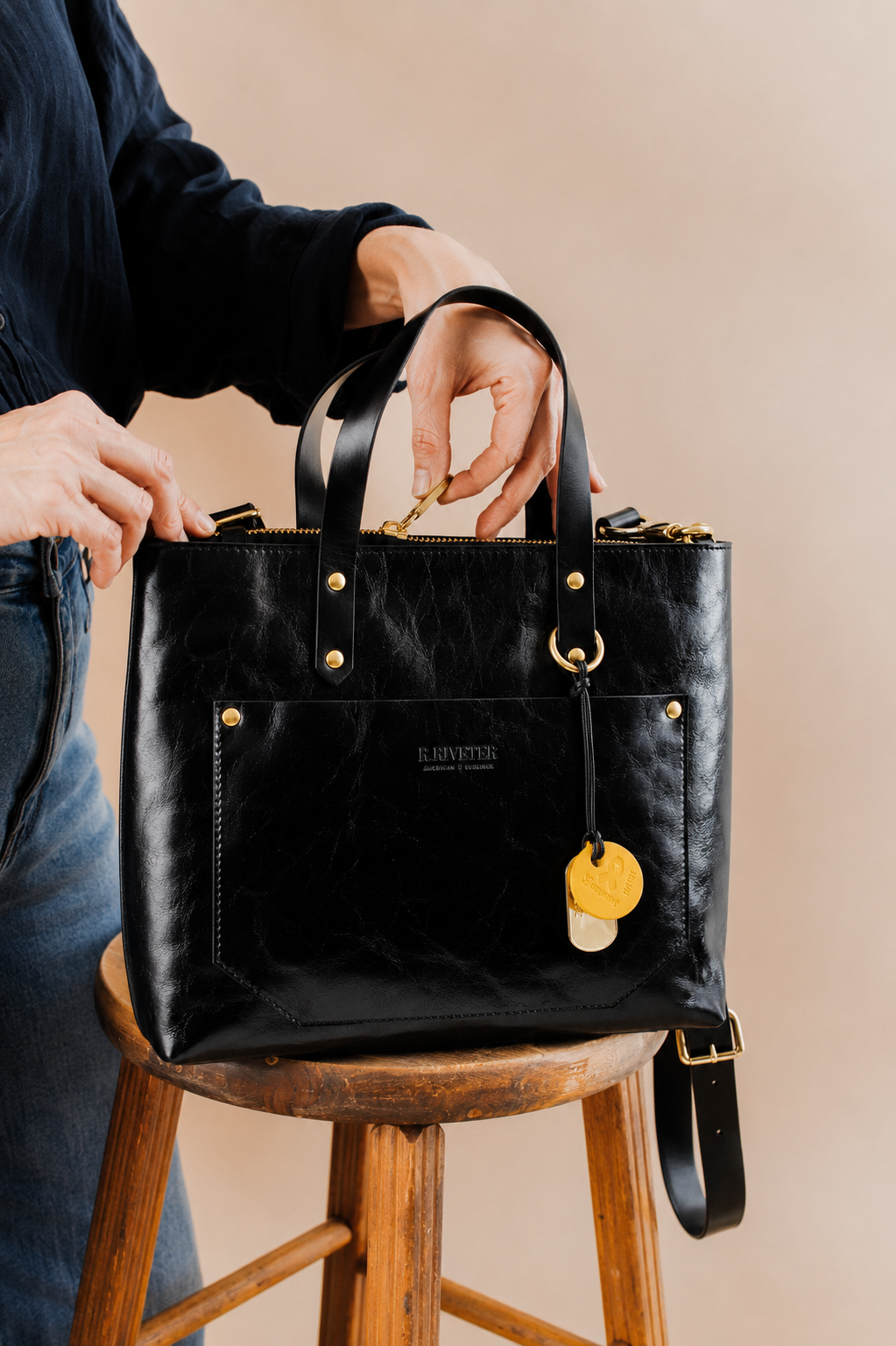 A black leather tote bag with gold hardware and a hanging yellow tag, held by a person seated on a wooden stool against a warm beige background.