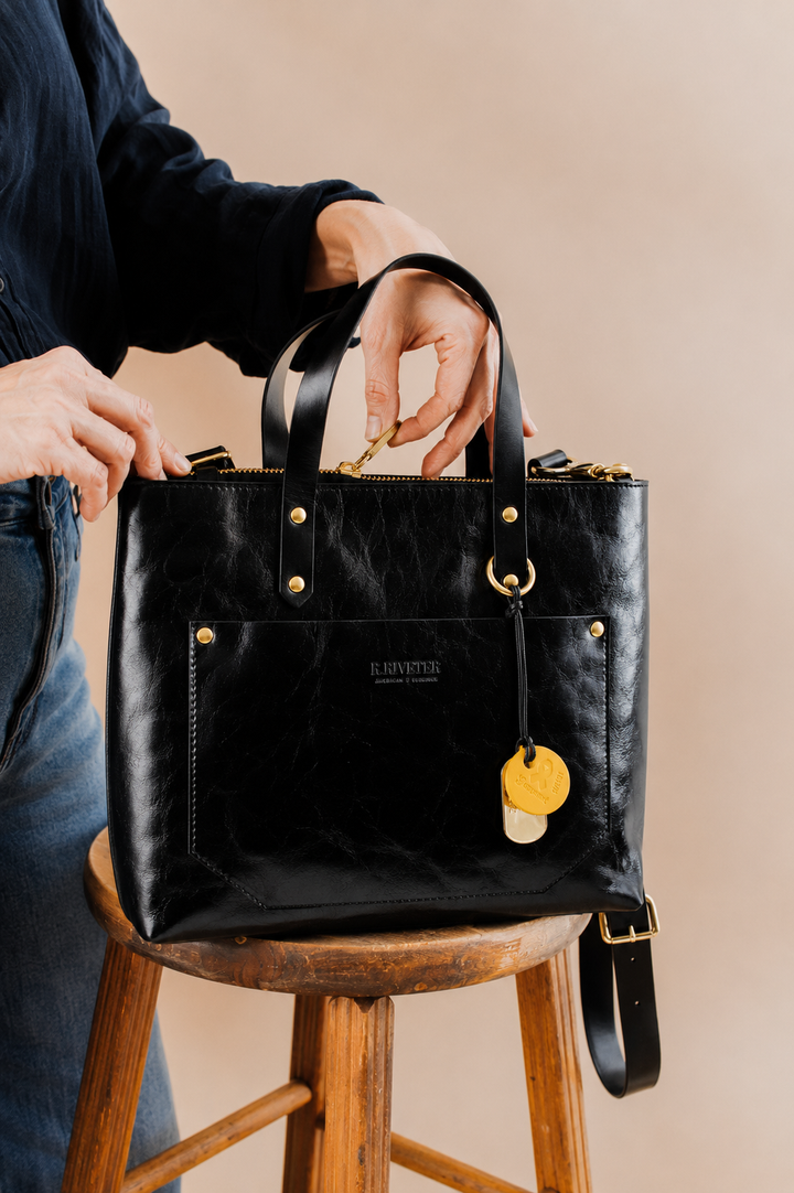 A black leather tote bag with gold hardware and a hanging yellow tag, held by a person seated on a wooden stool against a warm beige background.