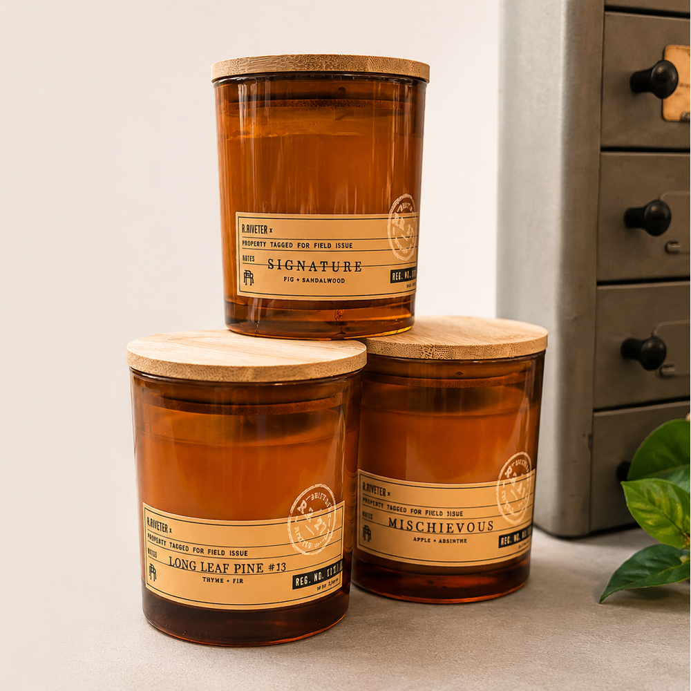 Three amber glass jar candles with light wood lids are arranged in a small stack on a tabletop. The labels read 'SIGNATURE' and 'MISCHIEFUS,' with 'HAND LEAF PURE CIL' visible.