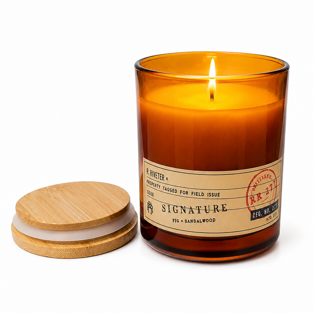 An amber glass jar candle with a lit wick and a separate round bamboo lid placed beside it on a clean white background. The front label reads Signature and includes scent text including barnwood.