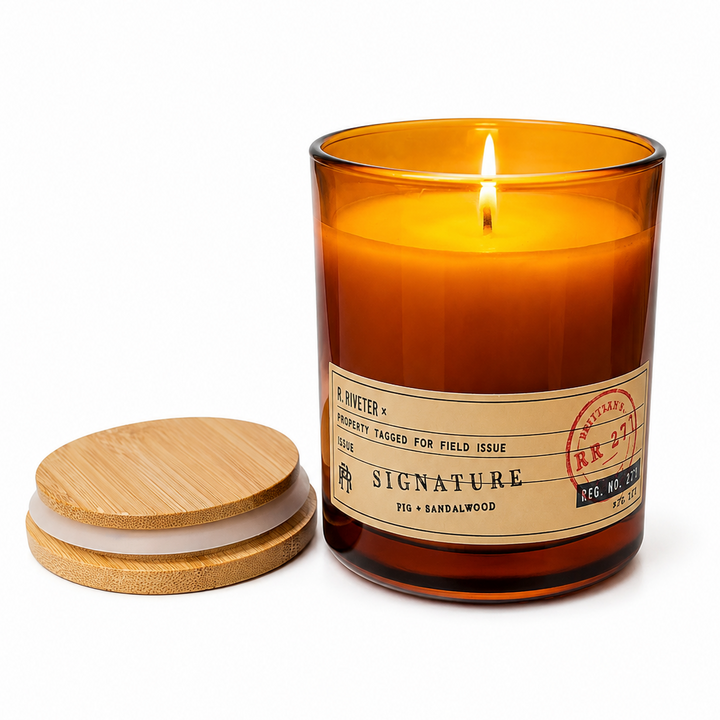 An amber glass jar candle with a lit wick and a separate round bamboo lid placed beside it on a clean white background. The front label reads Signature and includes scent text including barnwood.