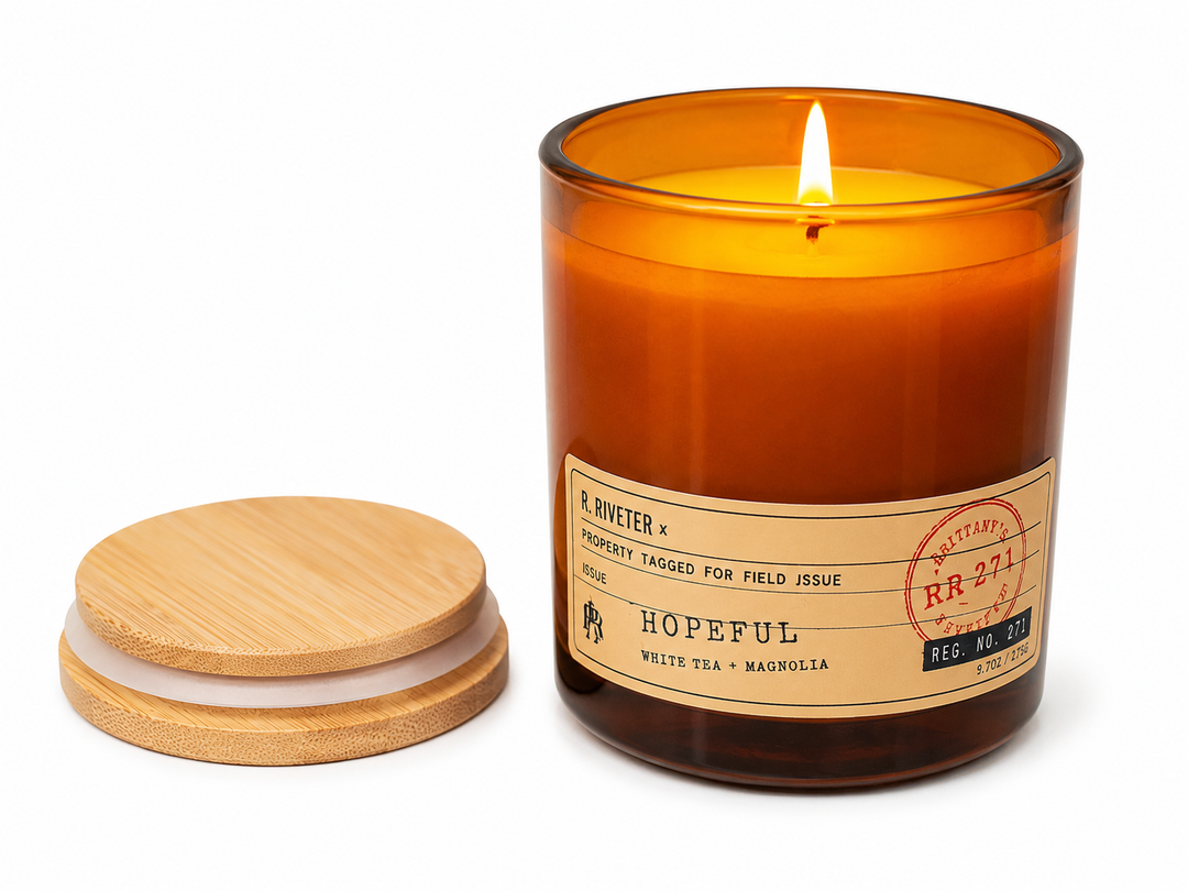 An amber glass jar candle with a lit wick and a beige paper label reading 'HOPEFUL' by R. Riveter, shown beside a bamboo lid on a white background.