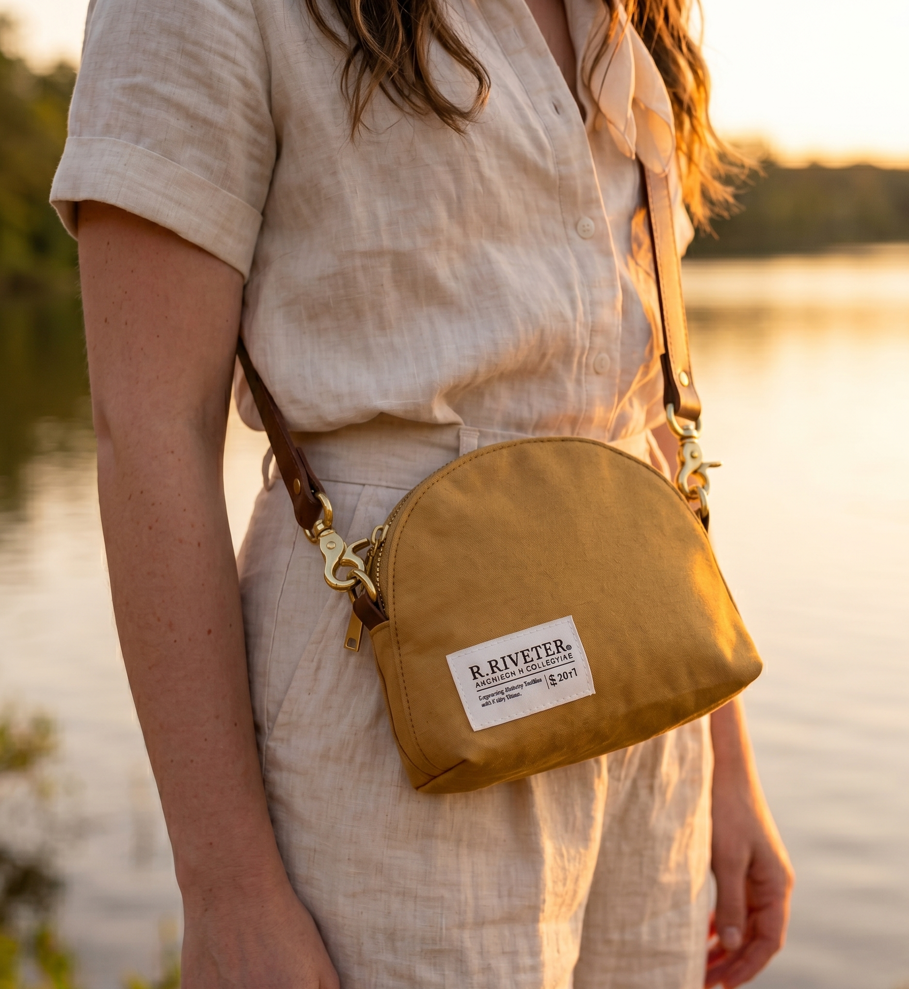 Person wearing a beige outfit with a brown leather bag by a body of water during sunset.